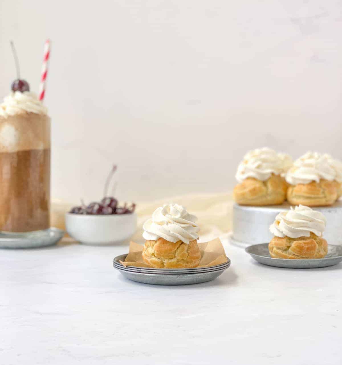 Root Beer Float Cream Puffs with whipped cream on top with more cream puffs, cherries, and a root beer float in the background