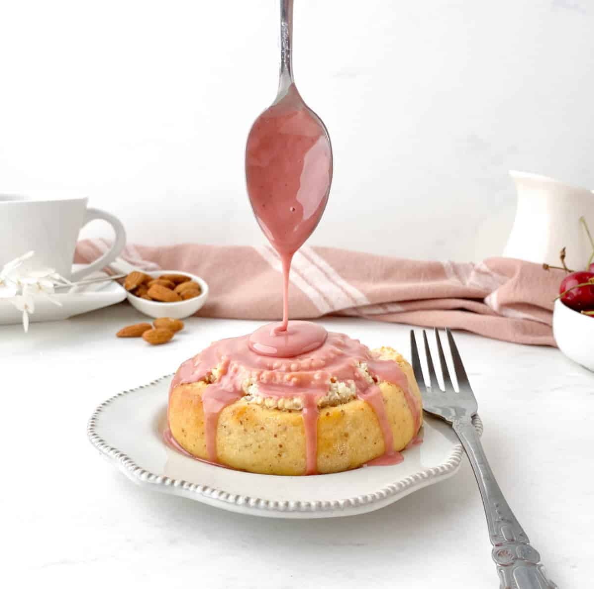 Pink glaze being drizzled on a Cherry Almond Sweet Roll on a white plate with a spoon 