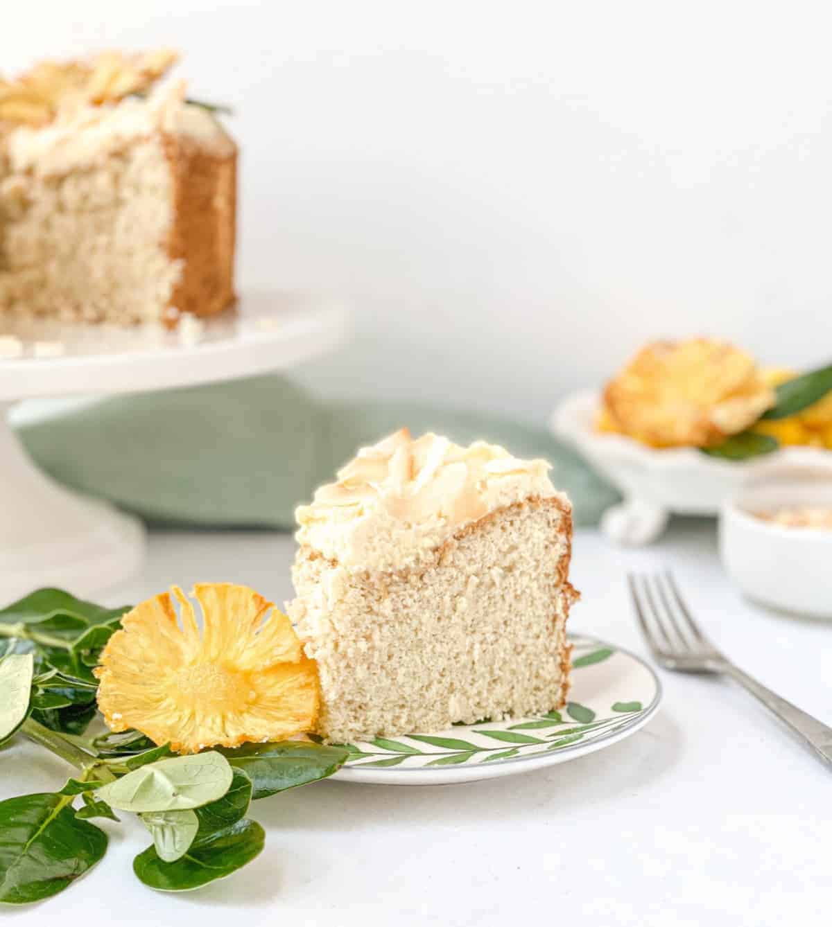Slice of Tropical Angel Food Cake on a plate with tropical leaves and pineapple slices nearby