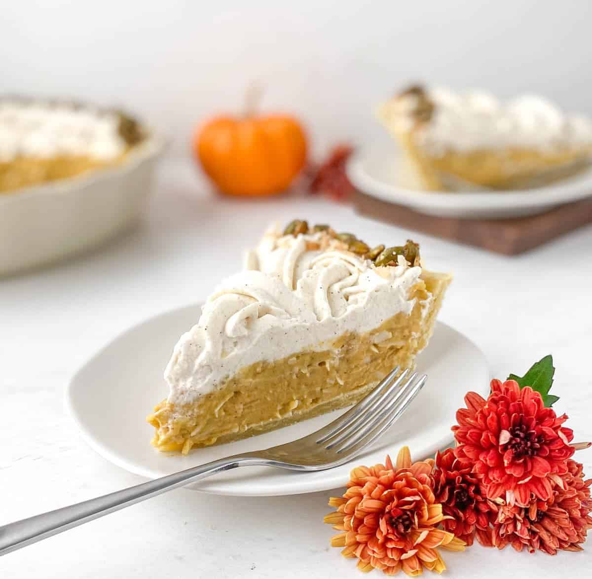 Slice of Pumpkin Coconut Cream Pie on a white plate with red mum flowers nearby