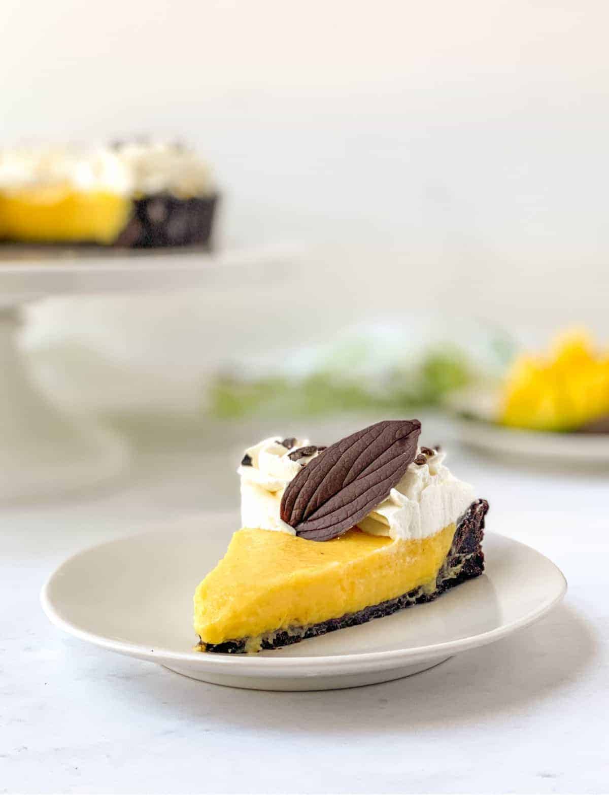 Slice of Chocolate Mango Tart on a white plate with a chocolate leaf on top, with the rest of the tart and mangos in the background