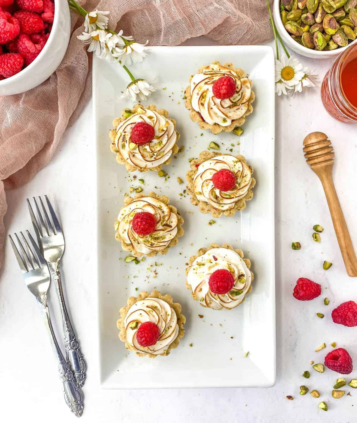 Overhead view of Raspberry Pistachio Tartlets on a rectangular white serving plate with raspberries, pistachios, and a jar of honey nearby