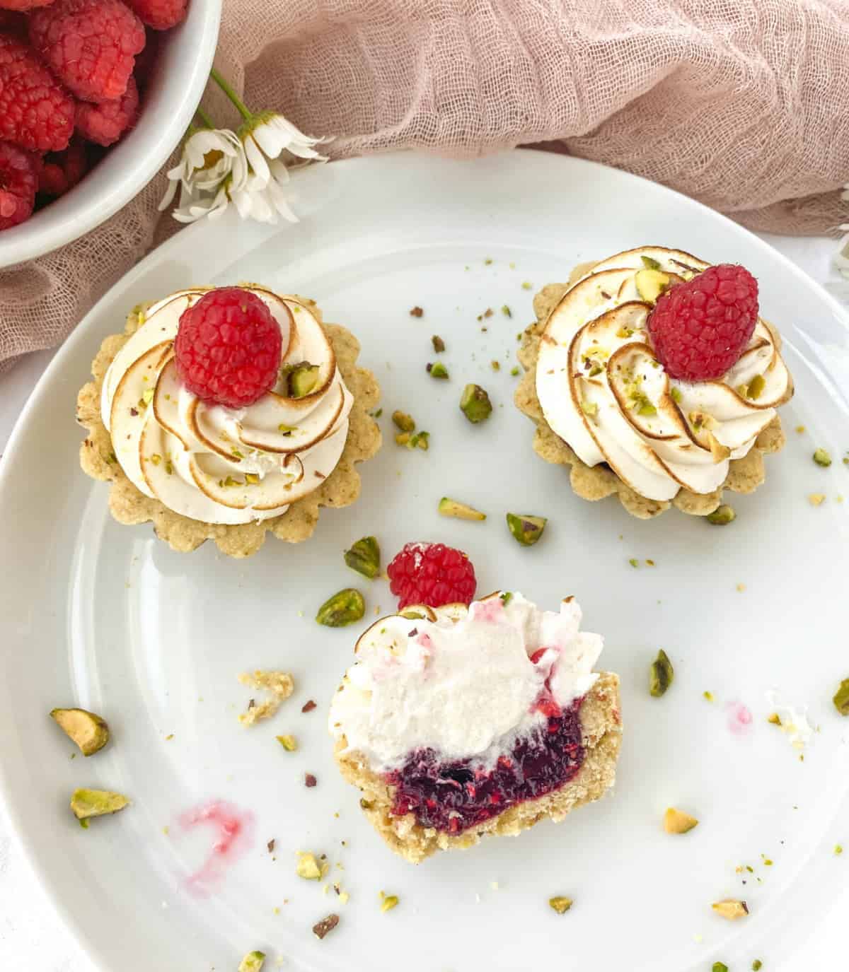Overhead view of three Raspberry Pistachio Tartlets on a white plate with chopped pistachios. One of the tartlets has been cut in half and is lying on its side.