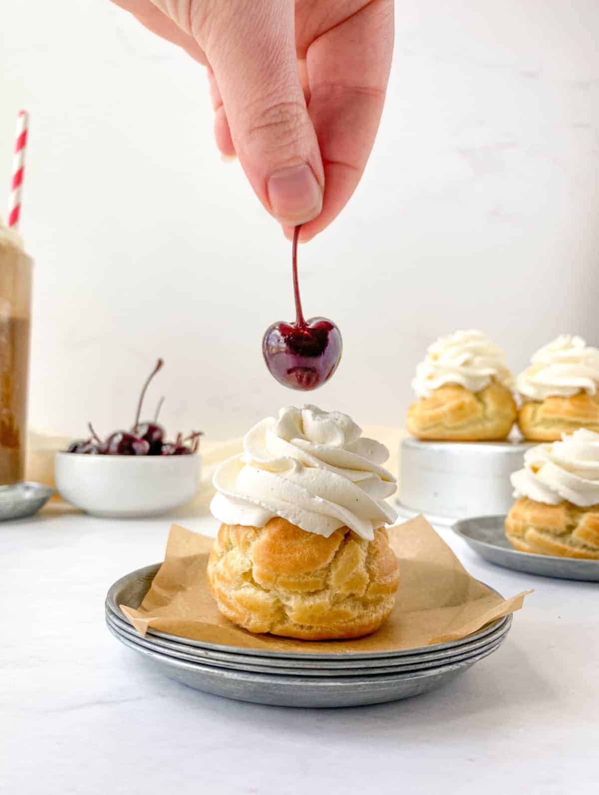 A hand placing a cherry on top of a Root Beer Float Cream Puff with more cream puffs and cherries in the background