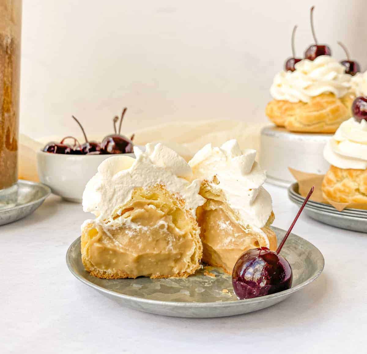 A Root Beer Float Cream Puff cut in half to see the root beer pastry cream on a silver plate with more cream puffs and cherries in the background