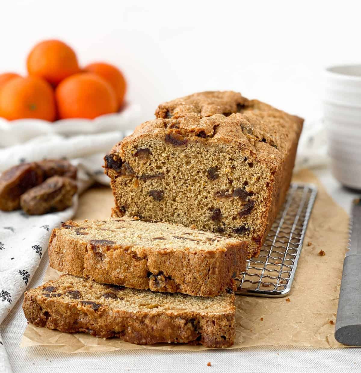 Sliced Date Bread on a cooling rack with dates and oranges nearby.