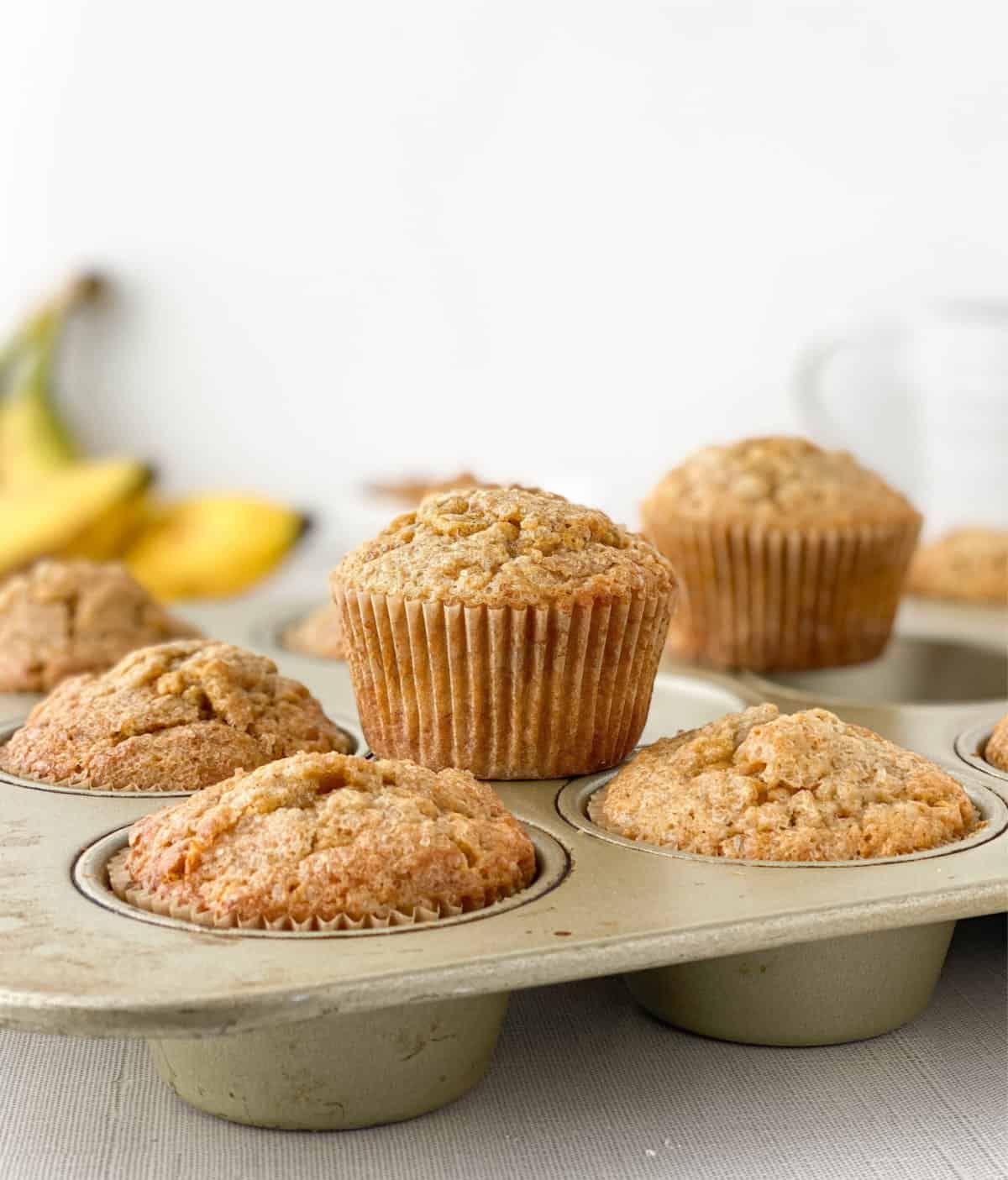 Butterscotch Banana Muffins in a muffin pan with bananas in the background.