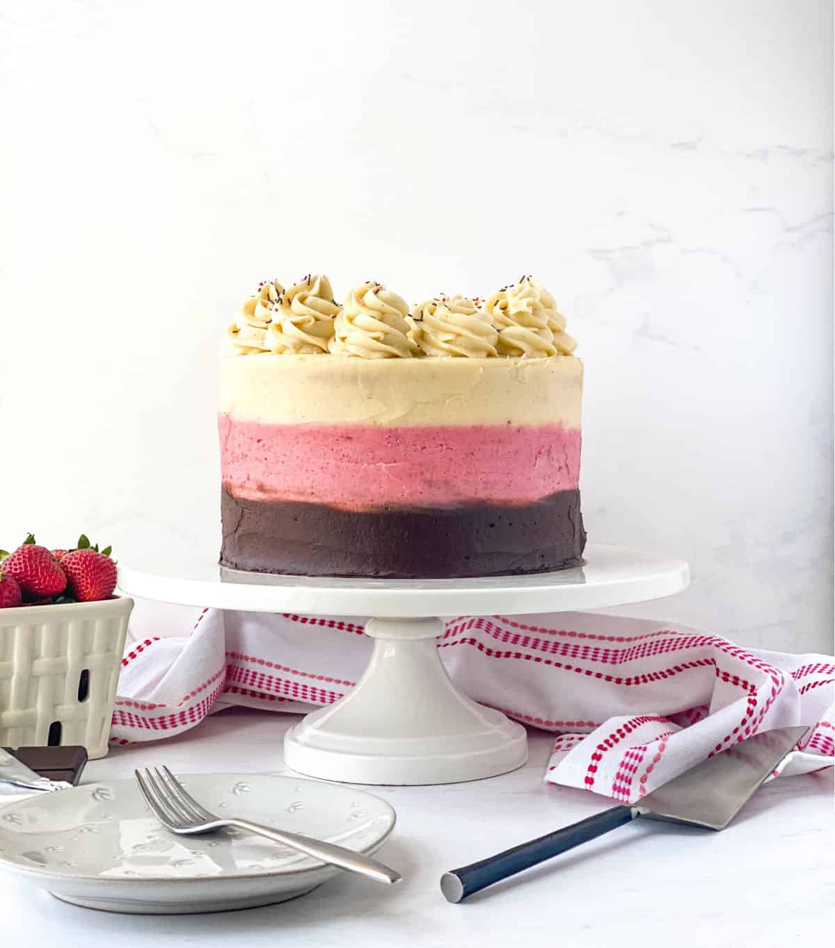 Neapolitan Cake on a white cake stand with strawberries nearby.