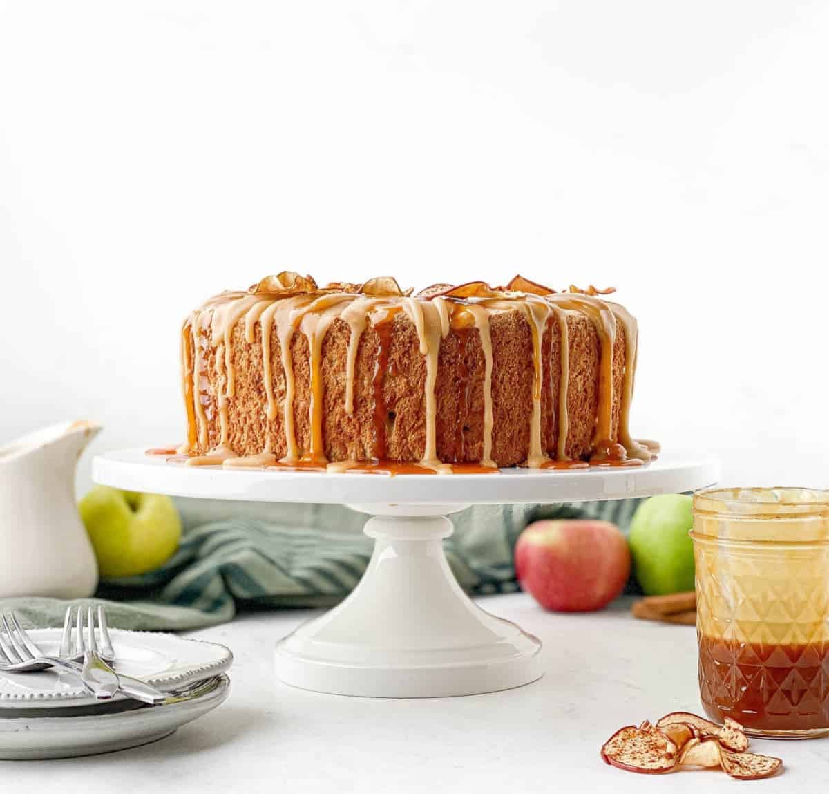 Caramel Apple Angel Food Cake on a white cake stand with apples, cinnamon sticks, and a jar of salted caramel sauce.
