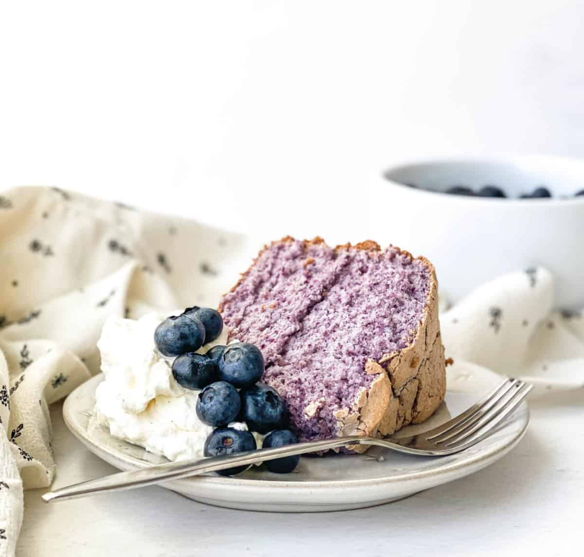 Slice of Blueberry Angel Food Cake with whipped cream and blueberries on a plate with a fork alongside.
