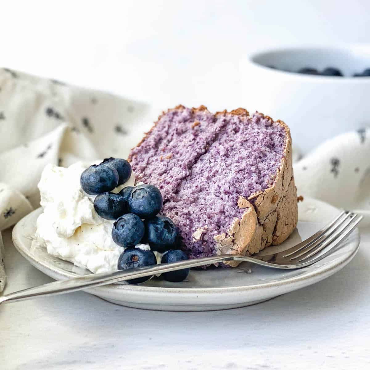 Slice of Blueberry Angel Food Cake on a plate with whipped cream and fresh blueberries.