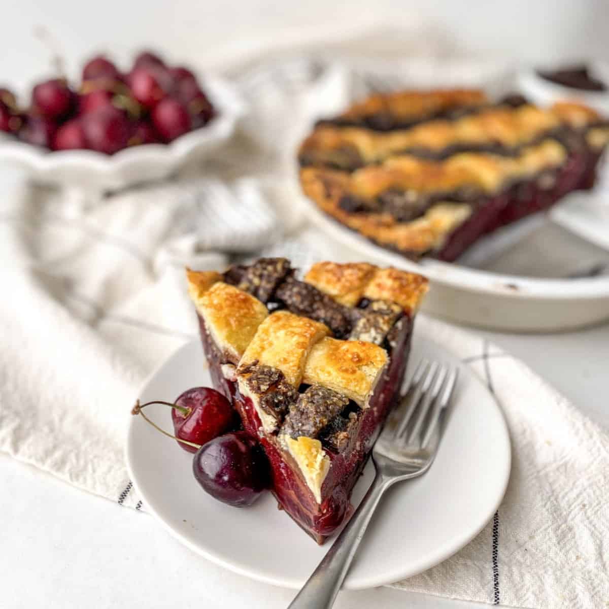 Slice of Chocolate Cherry Pie on a white plate with a bowl of cherries in the background.