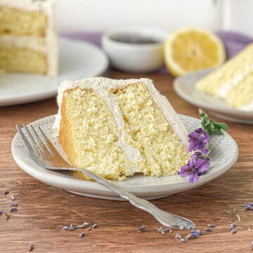 Slice of Lavender Lemon Cake on a white plate with lavender flowers alongside.