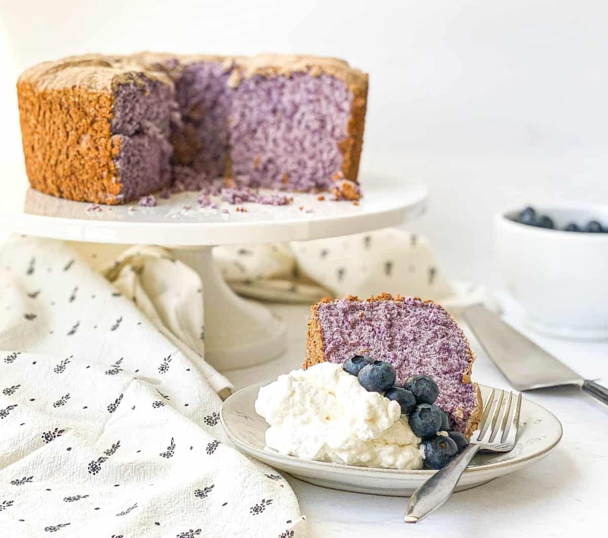 Slice of Blueberry Angel Food Cake on a white plate with whipped cream and fresh blueberries.