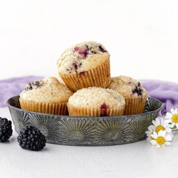 Tin of Blackberry Buttermilk Muffins with blackberries and chamomile flowers nearby.