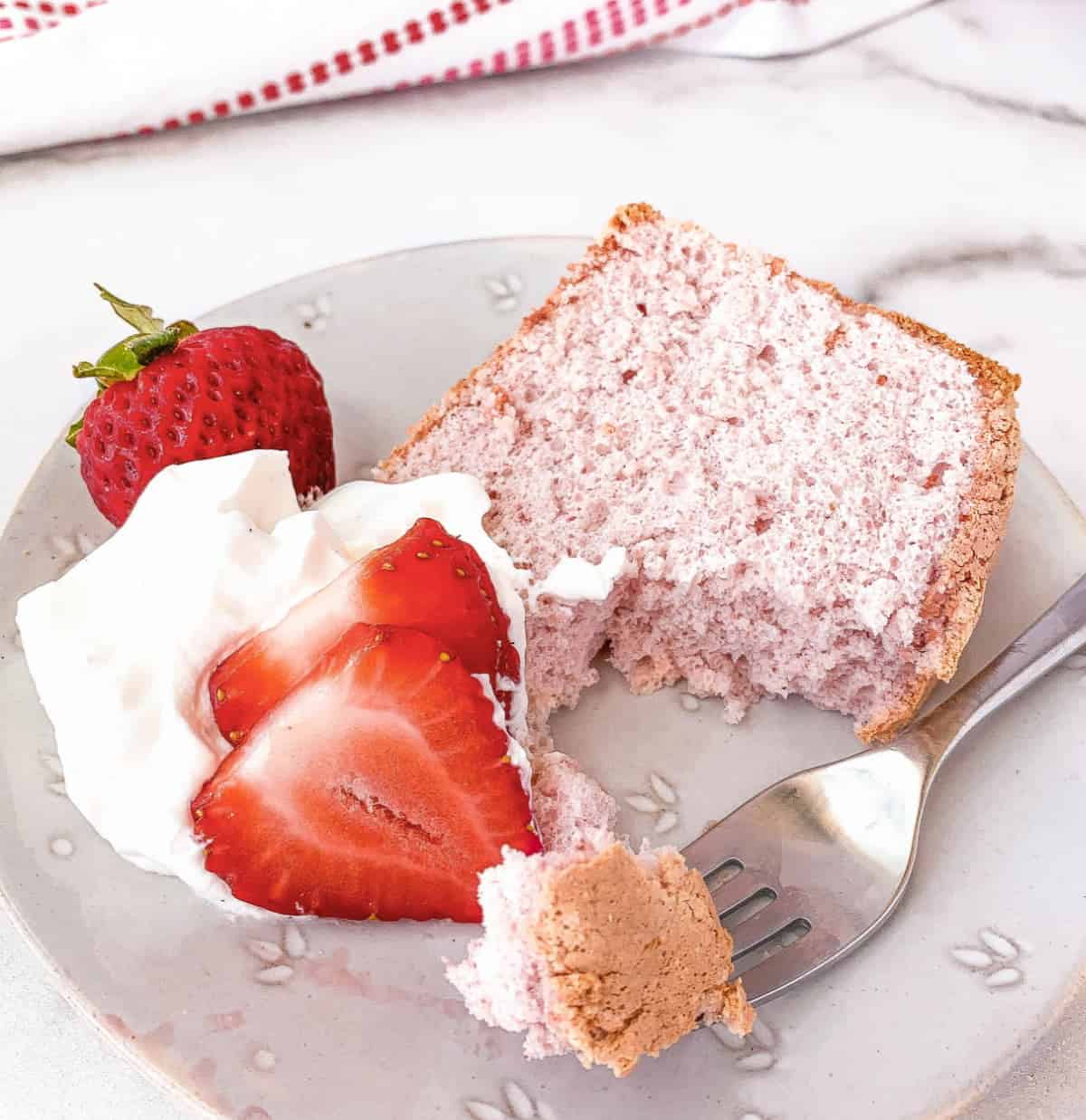 Partially eaten slice of Strawberry Angel Food Cake on a plate with whipped cream and fresh strawberries.