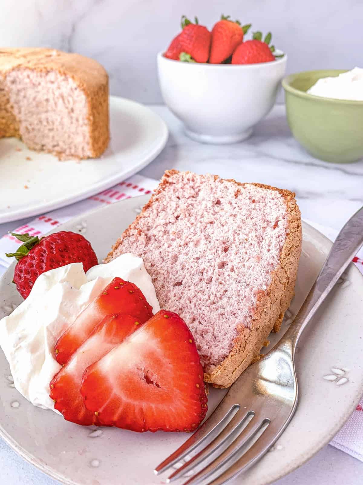 Slice of Strawberry Angel Food Cake on a plate with whipped cream and fresh strawberries.