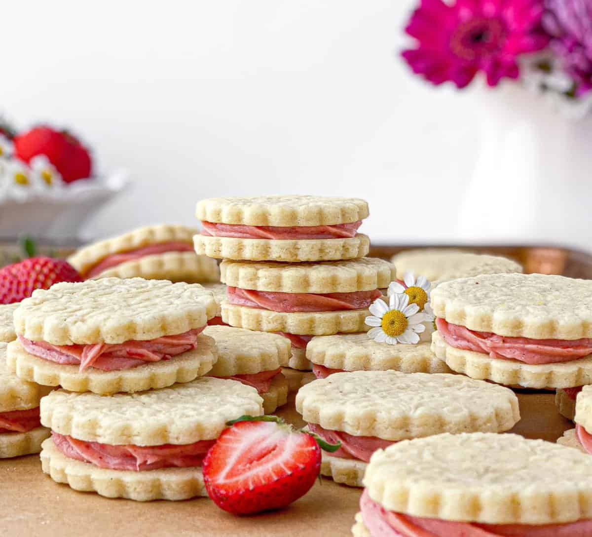 Strawberry Cheesecake Cookies stacked on a baking sheet with strawberries and small white flowers nearby.