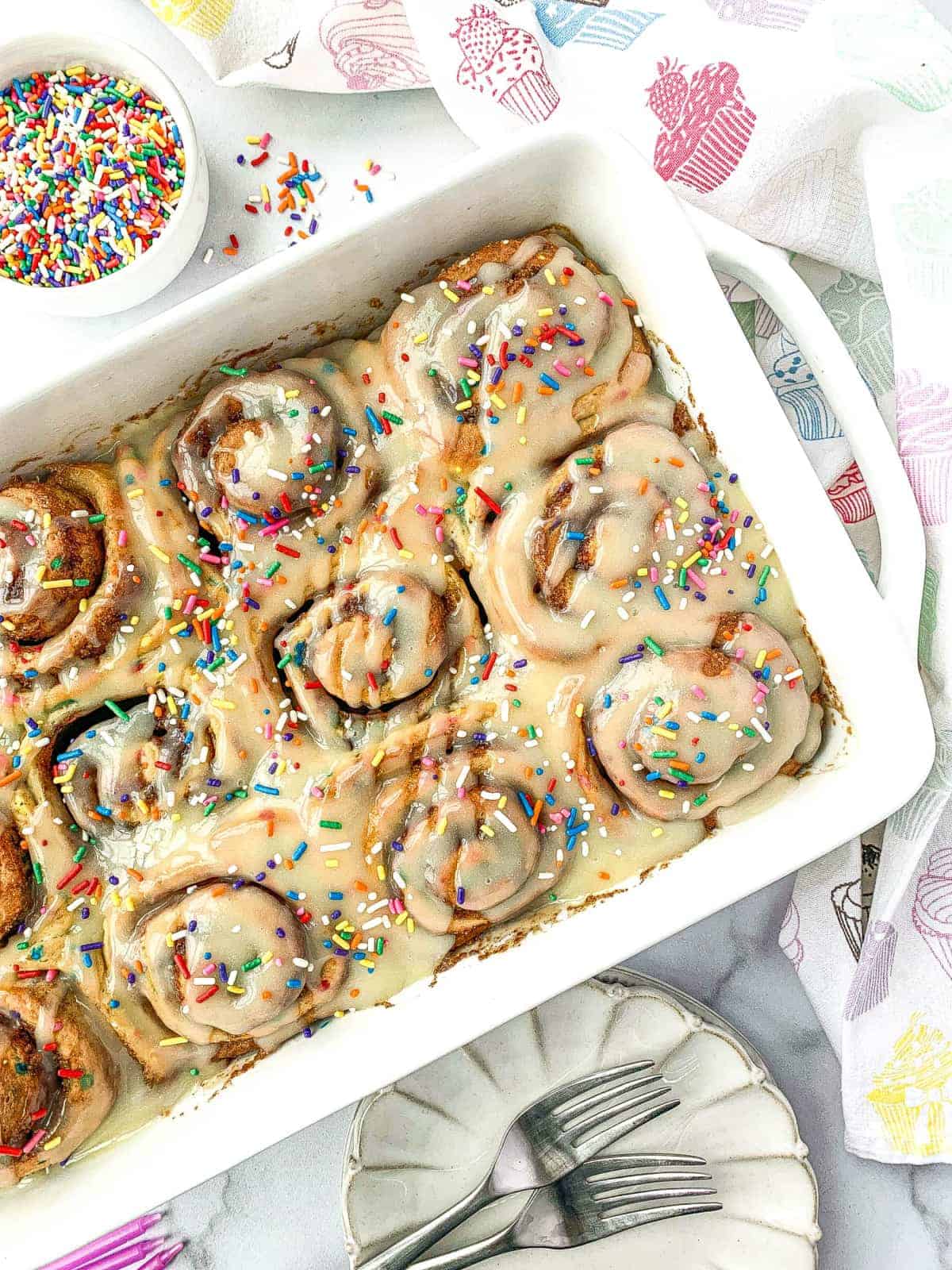 Funfetti Cinnamon Rolls in white pan with rainbow sprinkles nearby.