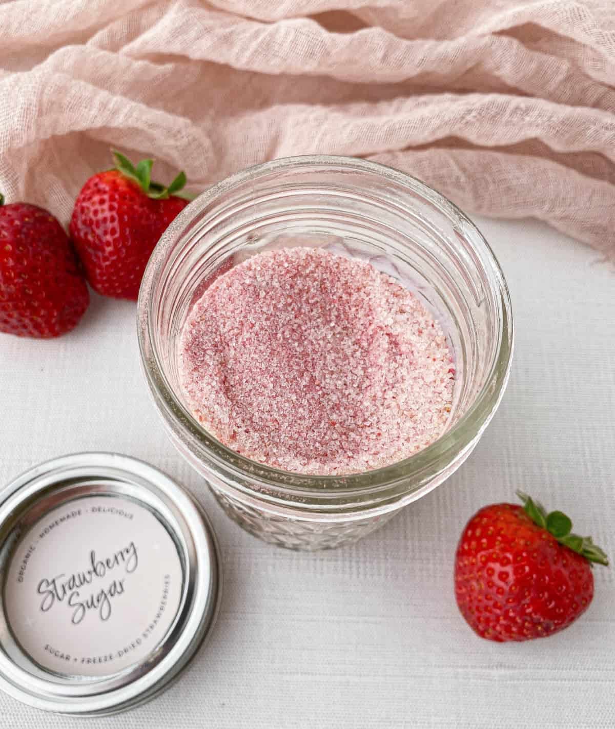 Open glass jar of Strawberry Sugar with fresh strawberries nearby.