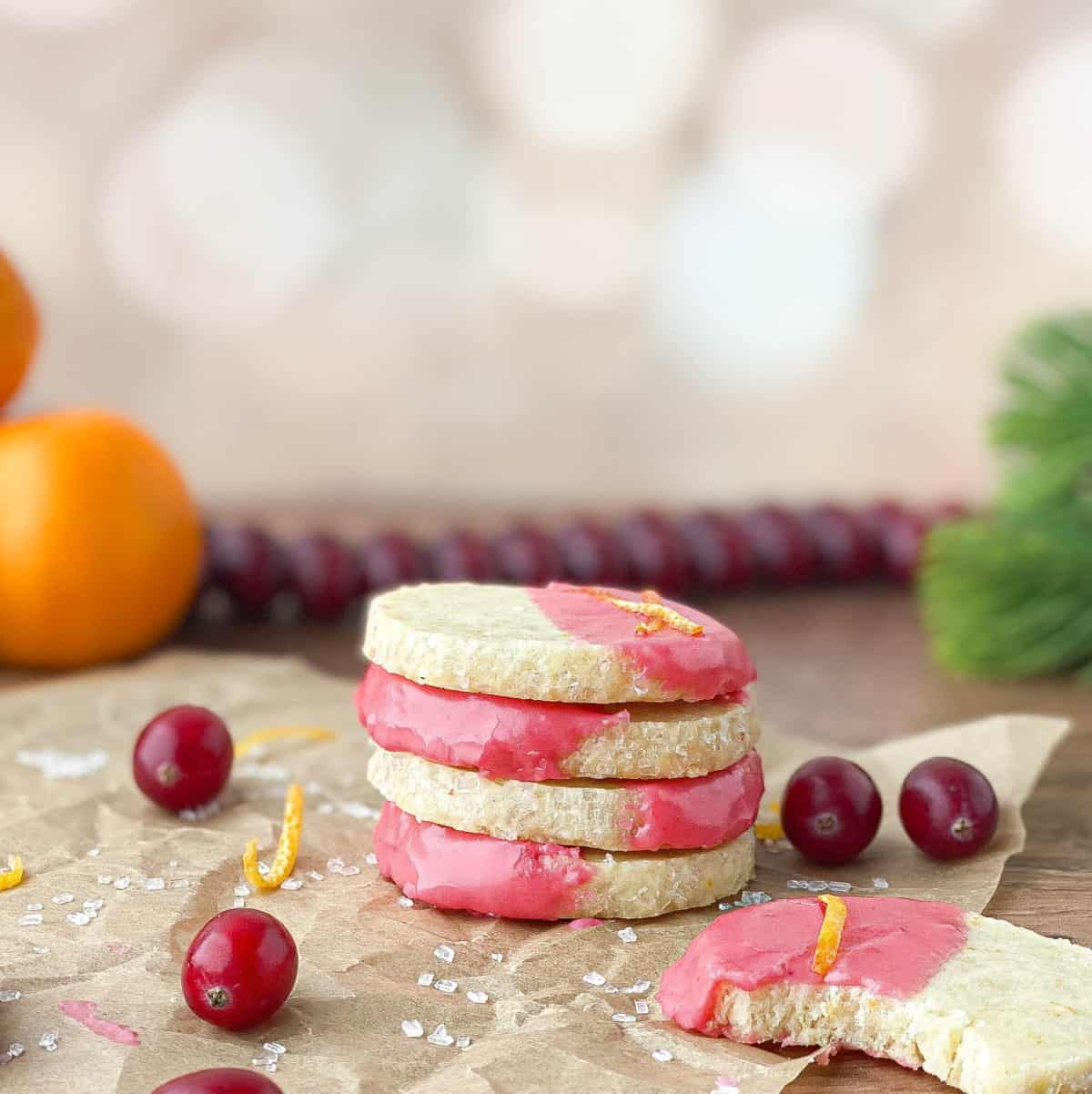 Stack of Orange Shortbread Cookies with Cranberry Glaze on parchment paper with fresh cranberries and oranges nearby.