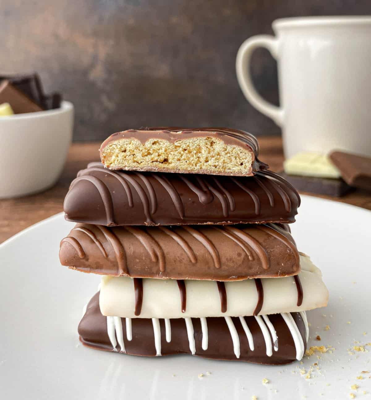 Stack of Chocolate Covered Graham Crackers with chocolate and coffee mug in the background.