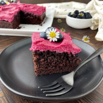 Slice of Blueberry Chocolate Cake on a black plate with fresh blueberries and a chamomile flower on top.