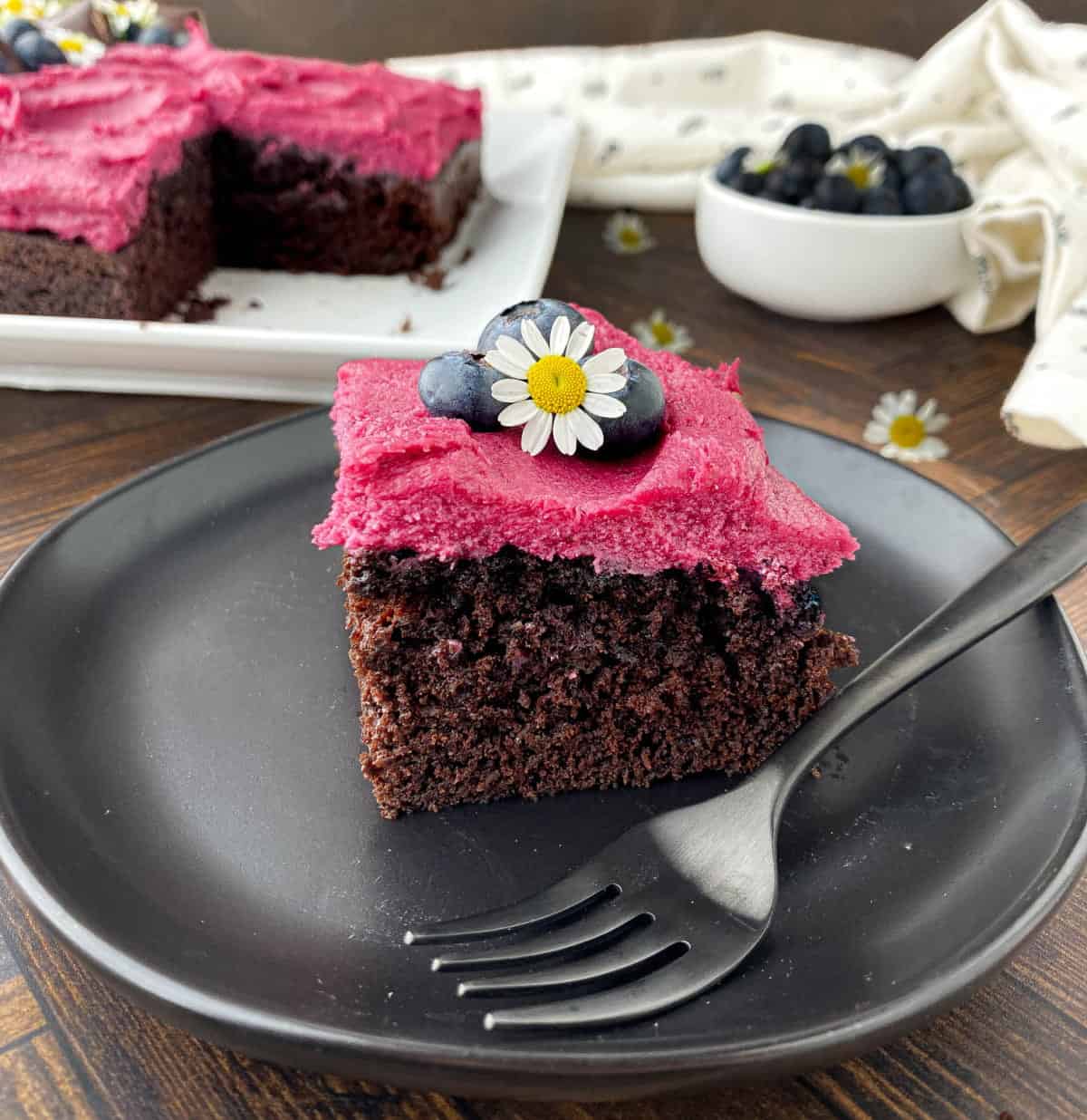 Slice of Blueberry Chocolate Cake on a black plate with fresh blueberries and a chamomile flower on top.
