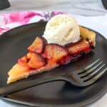Slice of Plum Tart with a scoop of vanilla ice cream on top on a black plate with a black fork alongside.