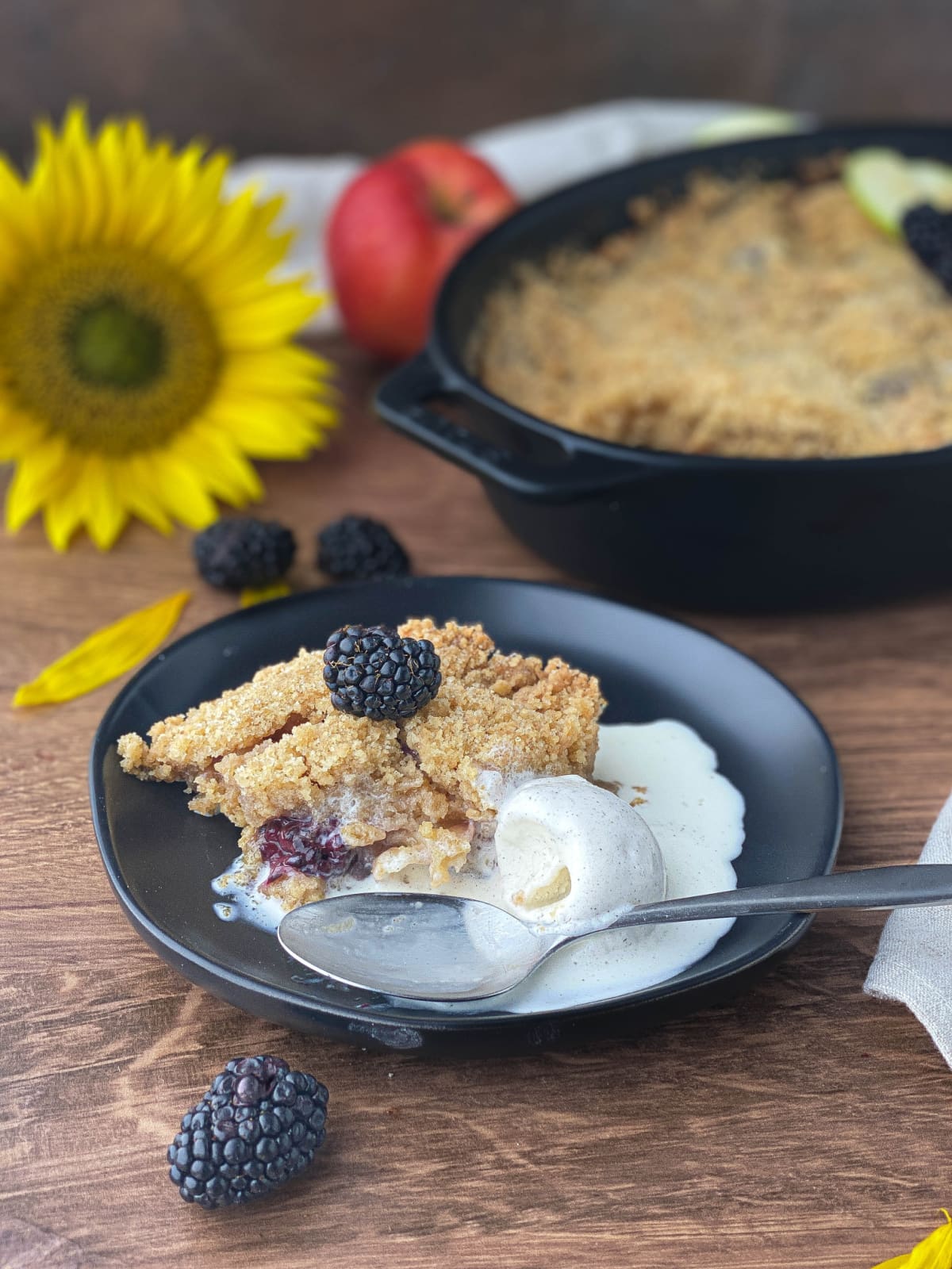 Blackberry and Apple Crumble on a black plate with a scoop of vanilla ice cream and a silver spoon.