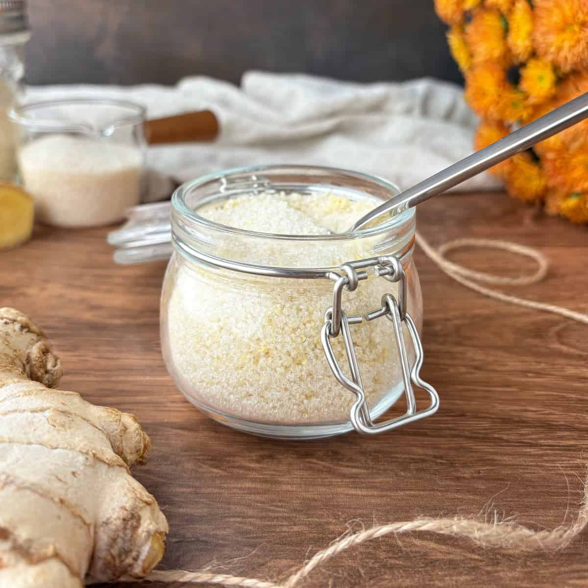 Small glass jar of Ginger Sugar surrounded by fresh ginger and orange mum flowers.