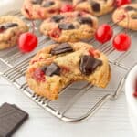 Closeup of Cherry Chocolate Chip Cookie with a bite missing on a silver cooling rack with maraschino cherries and more cookies in the background.