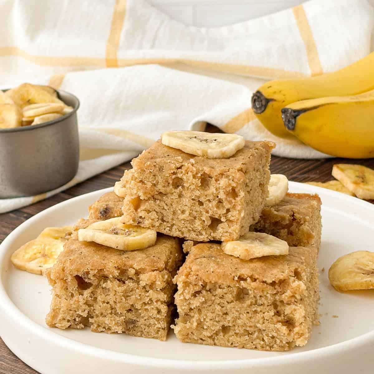 Three Banana Blondies stacked on a white plate topped with banana chips.