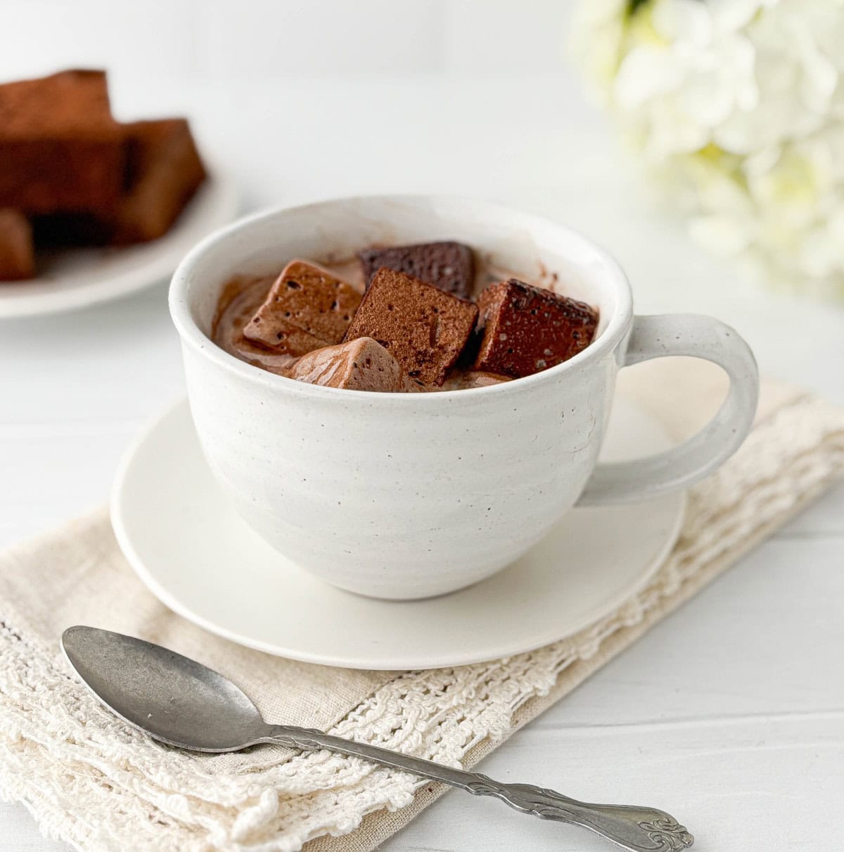 Chocolate Marshmallows in a white mug of hot chocolate on a white saucer with a silver spoon in the foreground.