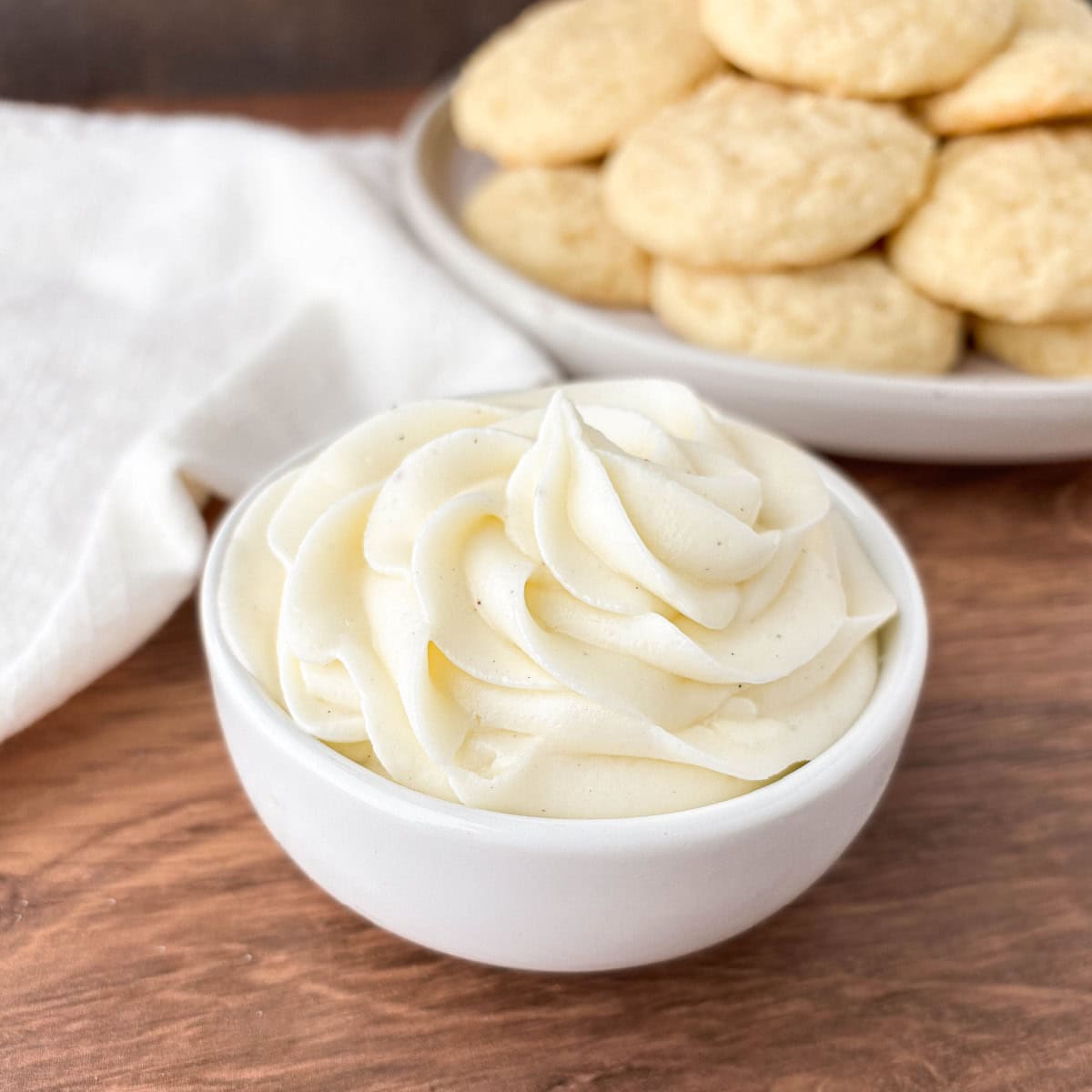 A small white bowl filled with Sour Cream Frosting with a plate of cookies in the background.