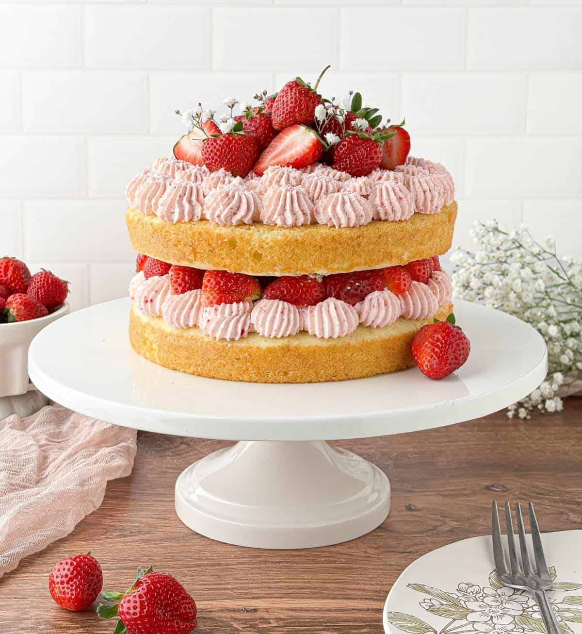 Strawberry Whipped Cream Cake on a white cake stand with fresh strawberries and white flowers in the background.