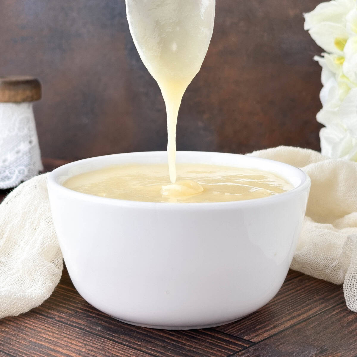 Silver spoon drizzling Cream Cheese Glaze into a small white bowl near an off-white towel.