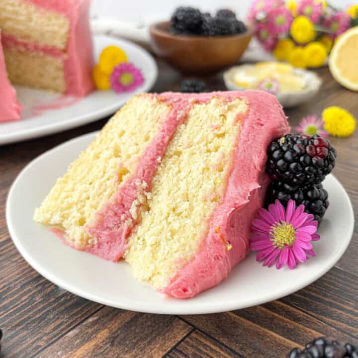 Slice of Lemon Blackberry Cake on a white plate with fresh blackberries and a small purple flower.
