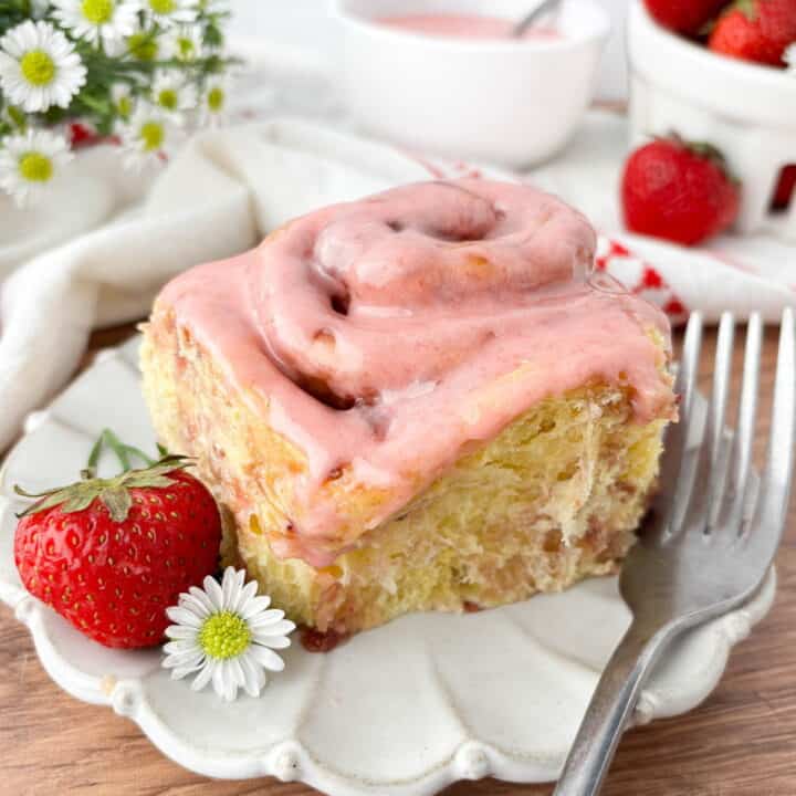 Closeup of Strawberry Cinnamon Roll topped with strawberry glaze on a white fluted plate with a strawberry, small white flower, and silver fork.