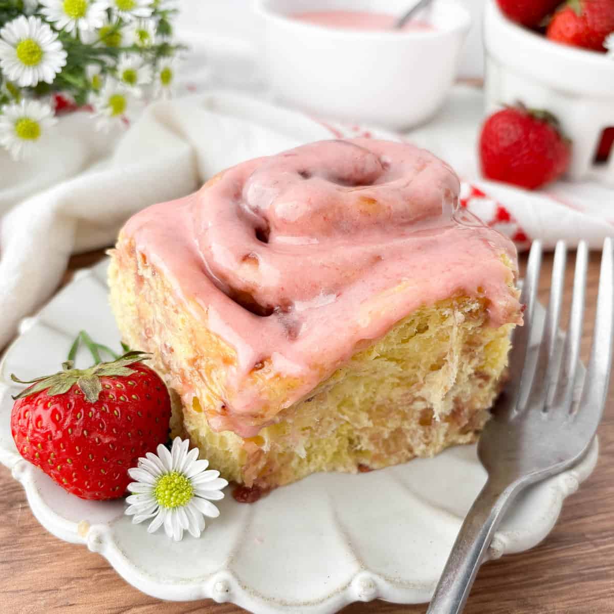Strawberry Cinnamon Roll on a white fluted plate with a fresh strawberry and silver fork.