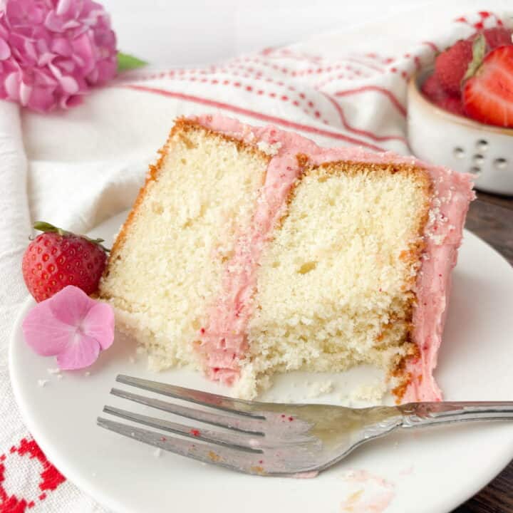 Partially eaten slice of Strawberry Vanilla Cake on a white plate with a silver fork, strawberry, and pink hydrangea flower.